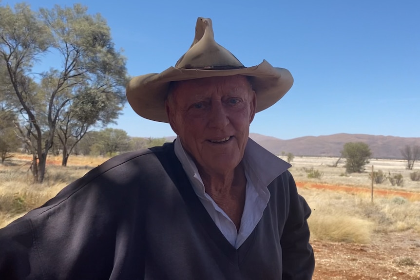An elderly man wearing a rugby jumper and a an old damaged broad brim hat smiles. There a striking ranges behind him.