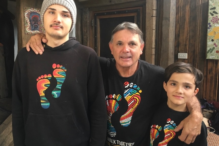 A tall Aboriginal man stands next to a shorter older Aboriginal man, who stands next to a young Aboriginal kid.