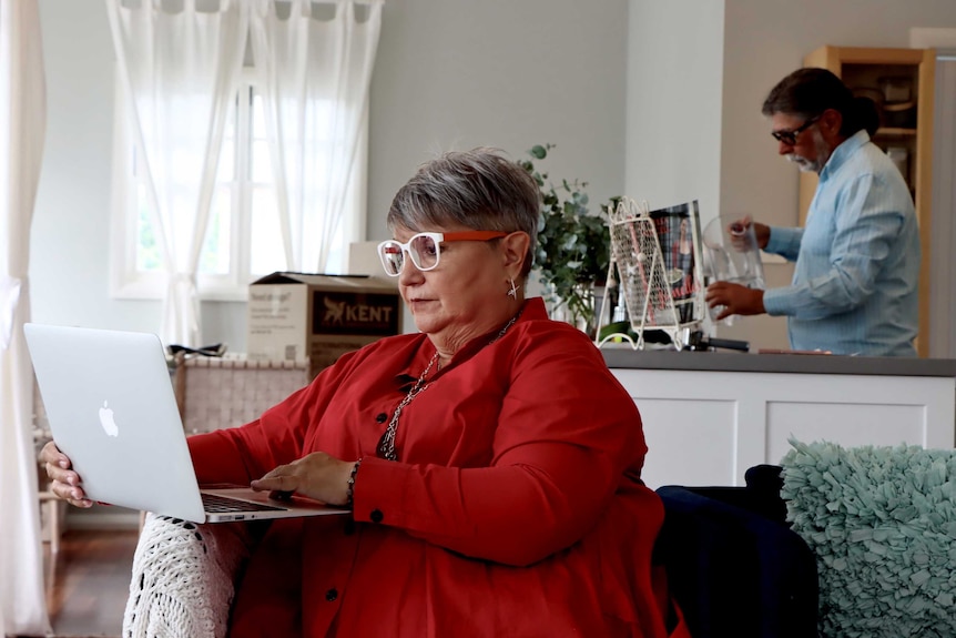 A woman seated on an armchair, looking at a laptop, with a man behind her pouring a glass of water.