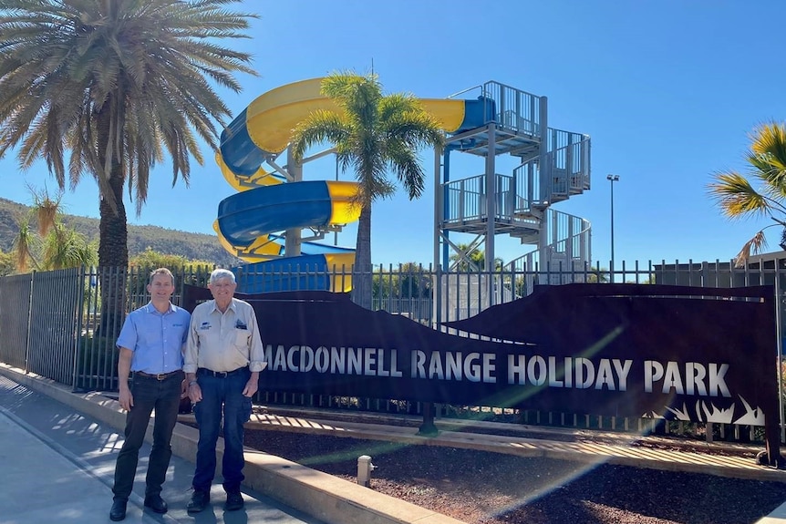 Two men stand in front of a sign that reads "MacDonnell Range Holiday Park".