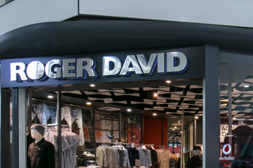 Six dummies dressed in menswear stand in the window of a store with a 'Roger David' sign