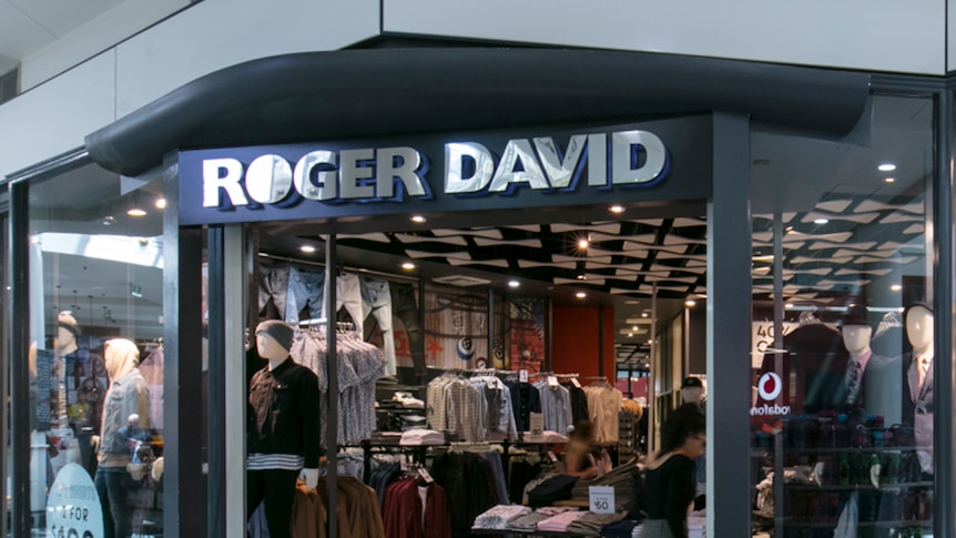 Six dummies dressed in menswear stand in the window of a store with a 'Roger David' sign