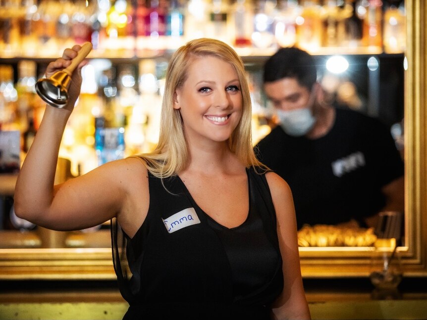 Lady standing in a bar holding a large bell, smiling