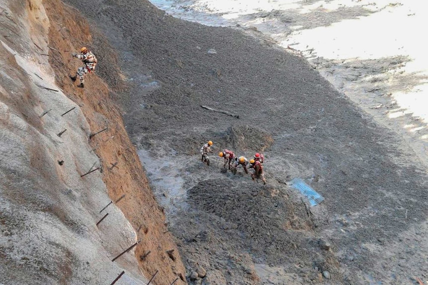 Five rescue personnel dig through mud and rock in an area smashed by surging water, while another rescuer abseils down to them.