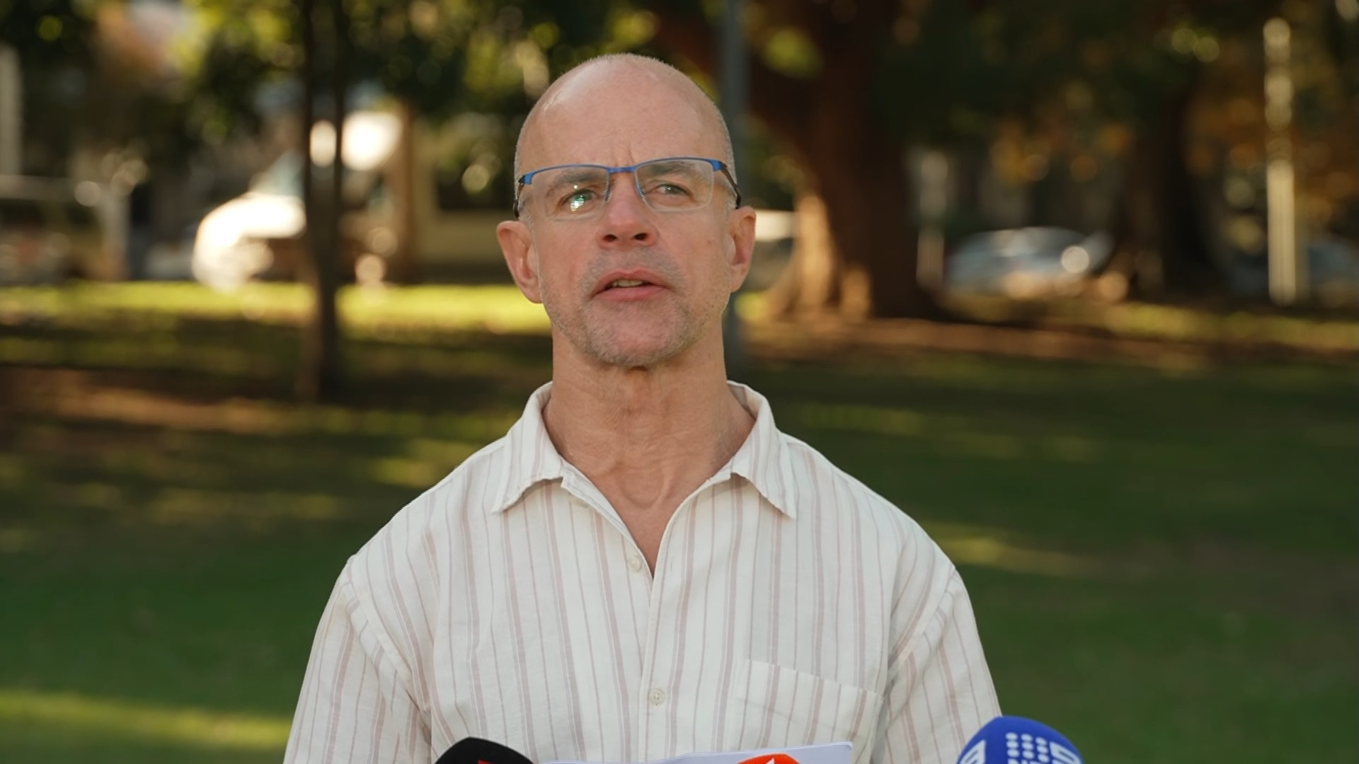 Con gafas y una camisa blanca a rayas, Nicholl habla con los medios de comunicación en un parque.