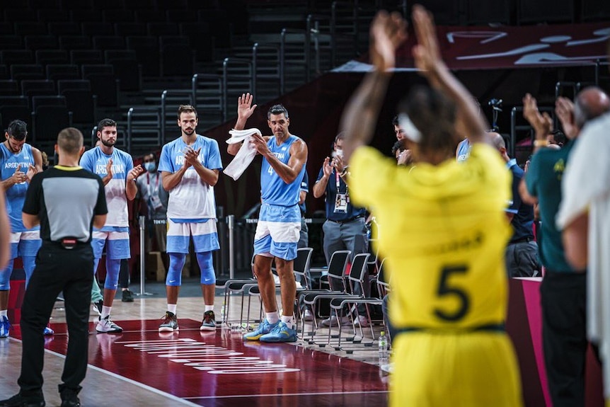 Luis Scola stands on a basketball court with his hand in the air as others stand and clap