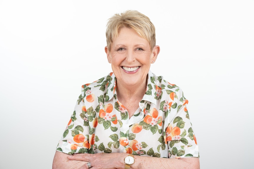 A woman in her late 70s with short blonde/grey hair in a shirt printed with peaches, sitting and smiling, white background