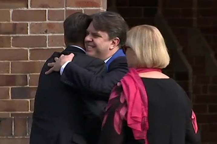 Two men hug while a woman looks on.