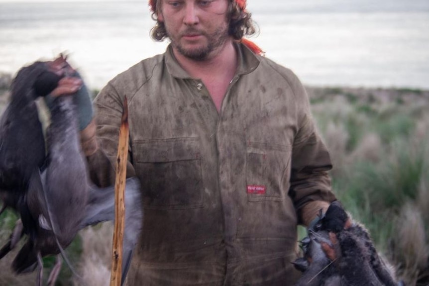 A man in overalls holds two dead birds.