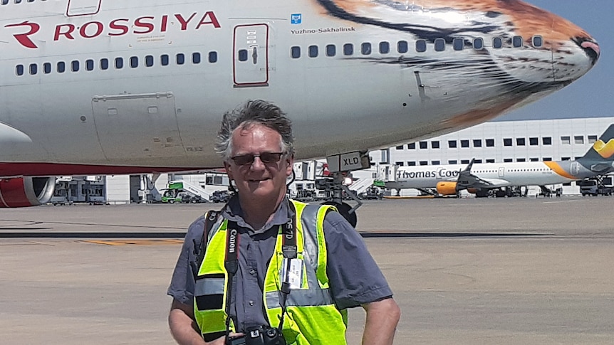 a man in a hi-vis vest with a camera around his neck stands on a tarmac with a plane behind