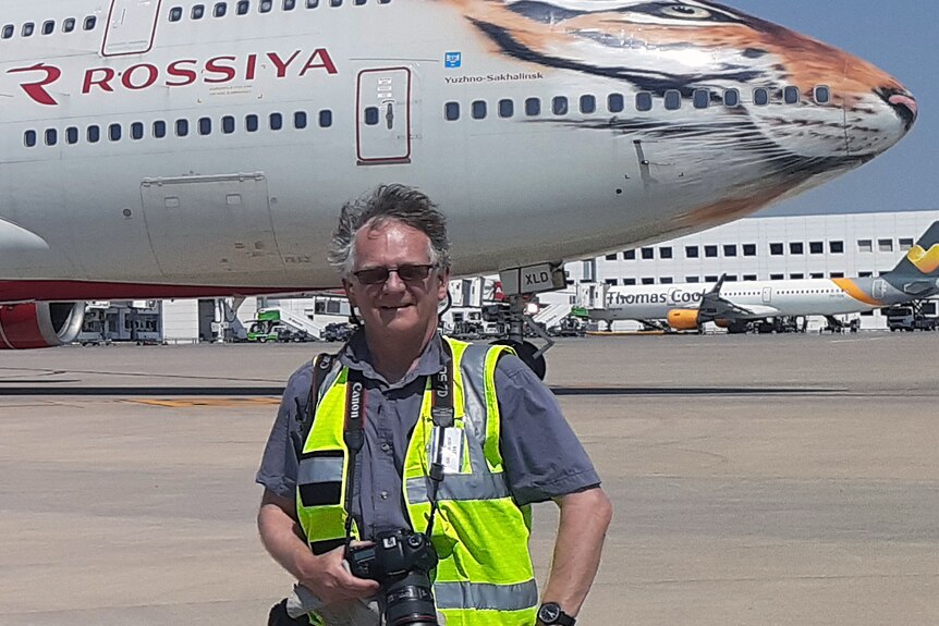a man in a hi-vis vest with a camera around his neck stands on a tarmac with a plane behind