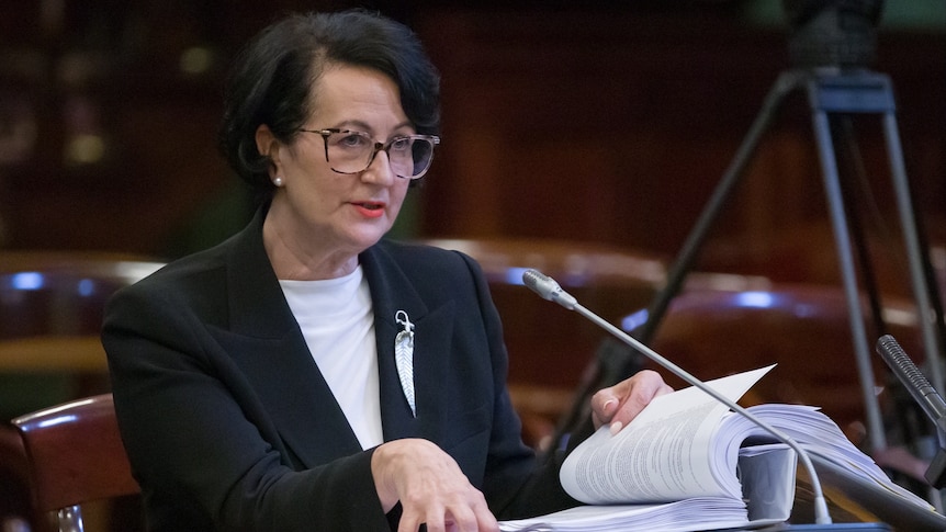 A woman wearing a black blazer, white top and glasses sits in front of a microphone. She is holding a binder of papers