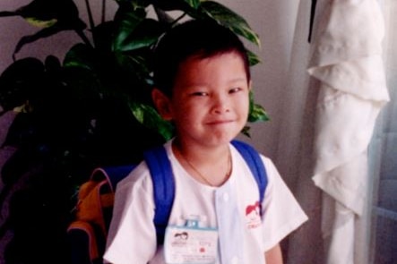 A young Asian boy looks to the camera and smiles with pursed lips. He stands in front of a plant and has a backpack on.