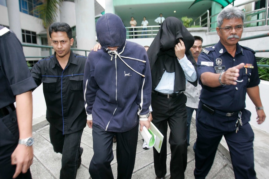 Police flank two men wearing hoodies over their faces outside court.