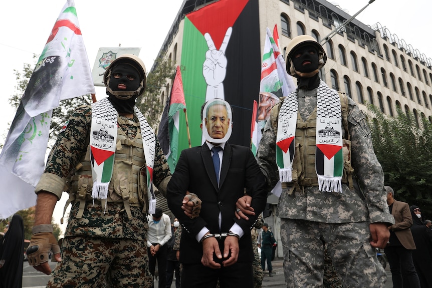 Two soldiers holding a man with a mask with Benjamin Netanyahu's face on it. 