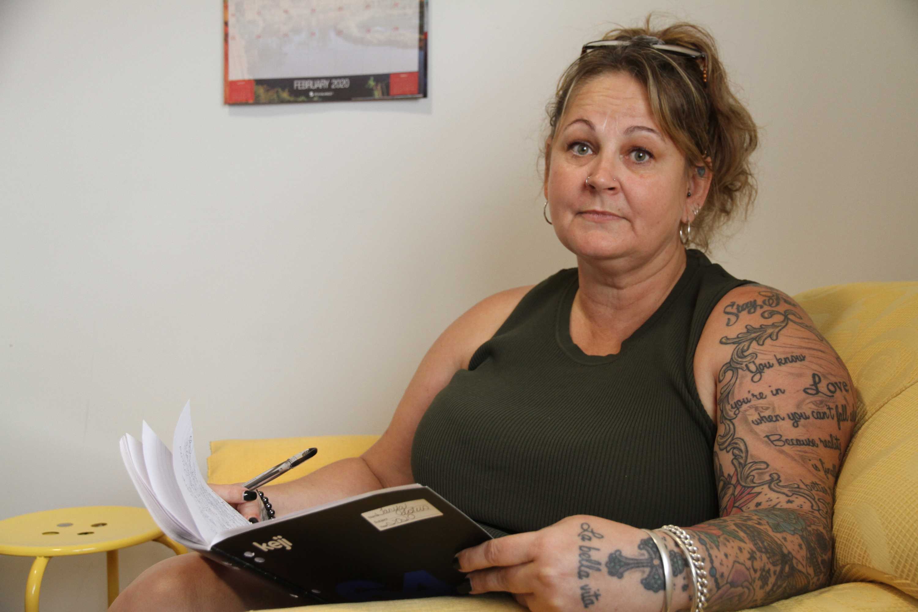 A middle-aged woman with tattoos holds a notebook and looks sadly at the camera.