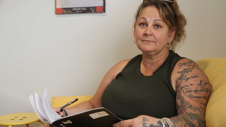 A middle-aged woman with tattoos holds a notebook and looks sadly at the camera.