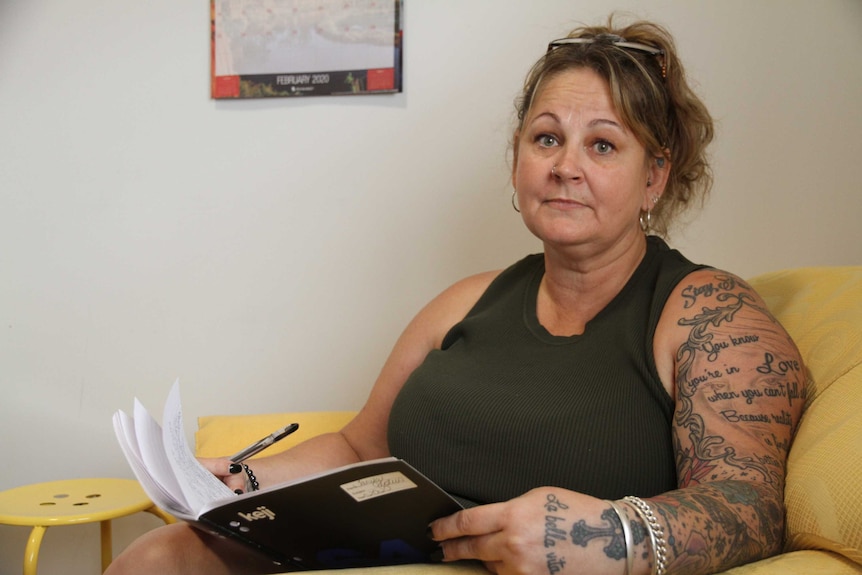 A middle-aged woman with tattoos holds a notebook and looks sadly at the camera.