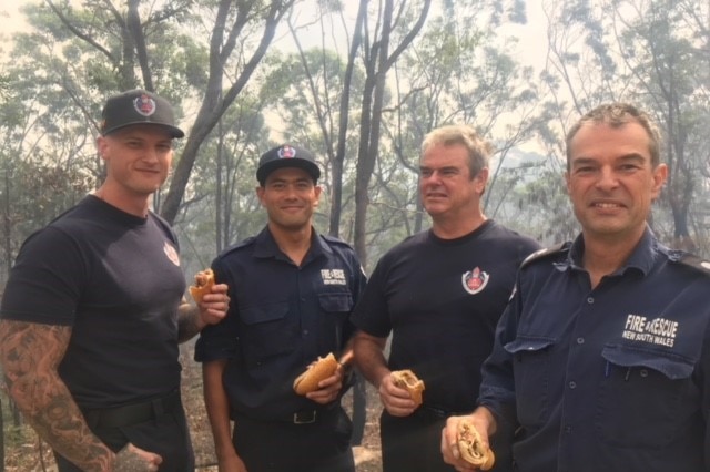 Four firefighter men holding sausage sandwiches.