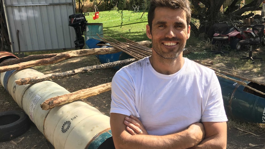 A smiling Mike Atkinson dressed in a white t-shirt stands in front of a home-made raft in a back yard.