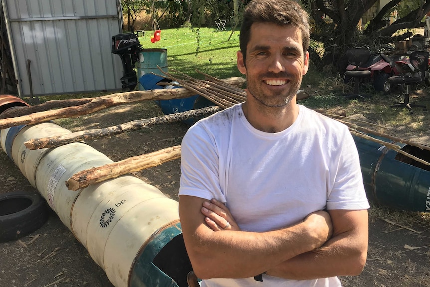 A smiling Mike Atkinson dressed in a white t-shirt stands in front of a home-made raft in a back yard.