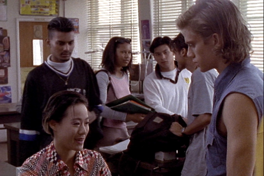 A group of teenagers stand around in a classroom talking