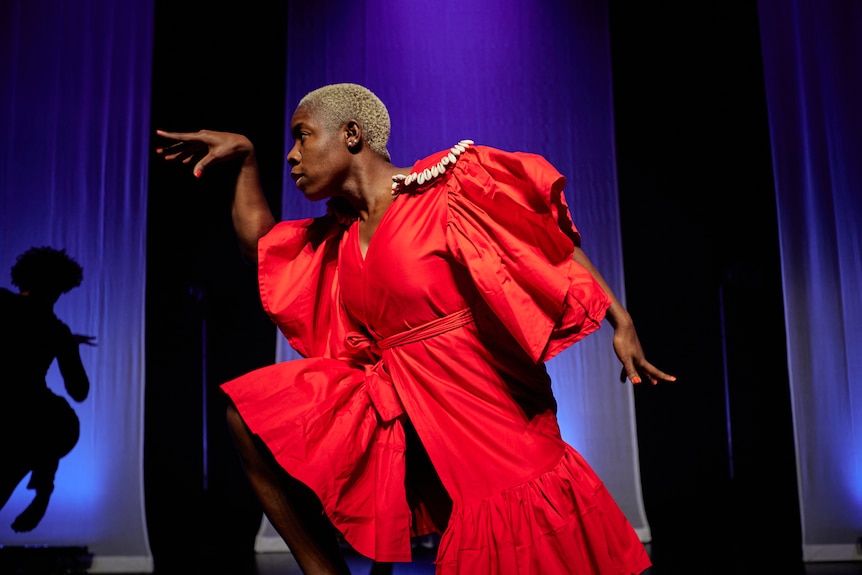 A Black woman, a dancer, in a red dress with puffy sleeves, poses on stage, one arm reaching behind her, the other in front