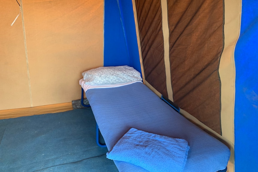 stretcher bed with blue blanket and floral pillow inside canvas tent with cream, brown and blue walls
