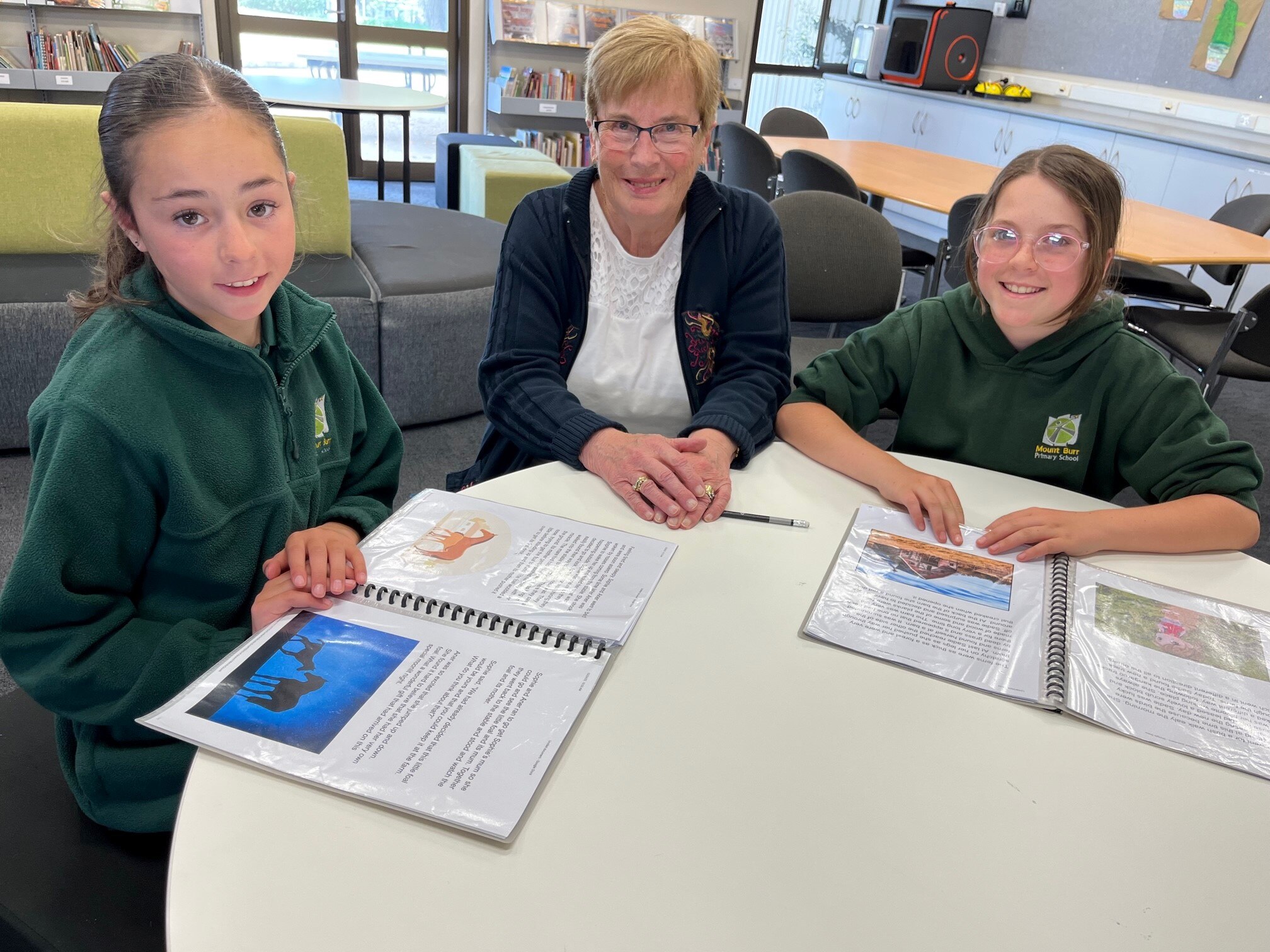Woman sitting between two girls in green school uniforms at small desk in a classroom.