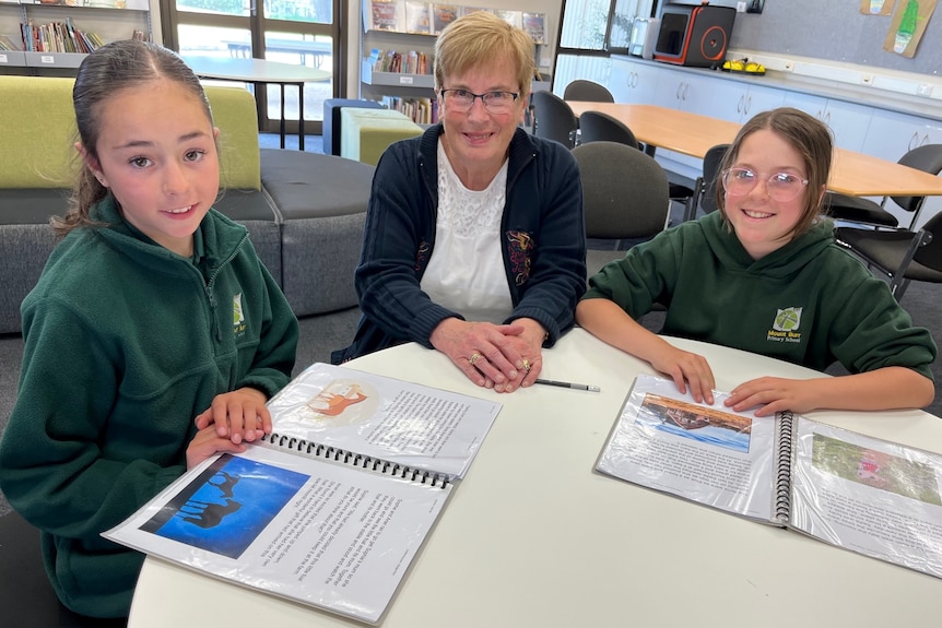 Woman sitting between two girls in green school uniforms at small desk in a classroom.