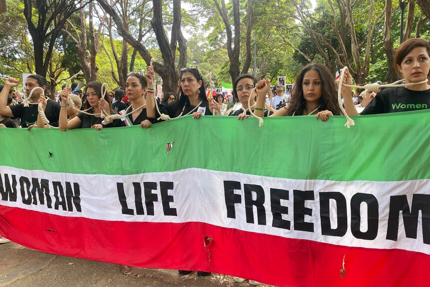 women hold nooses around their necks and a banner in the colours of the Iranian flag with woman life freedom written on it