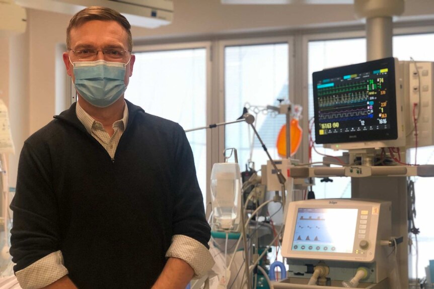 Dr Stephen Warrilow wears a facemask and stands in front of a machine in an intensive care unit.