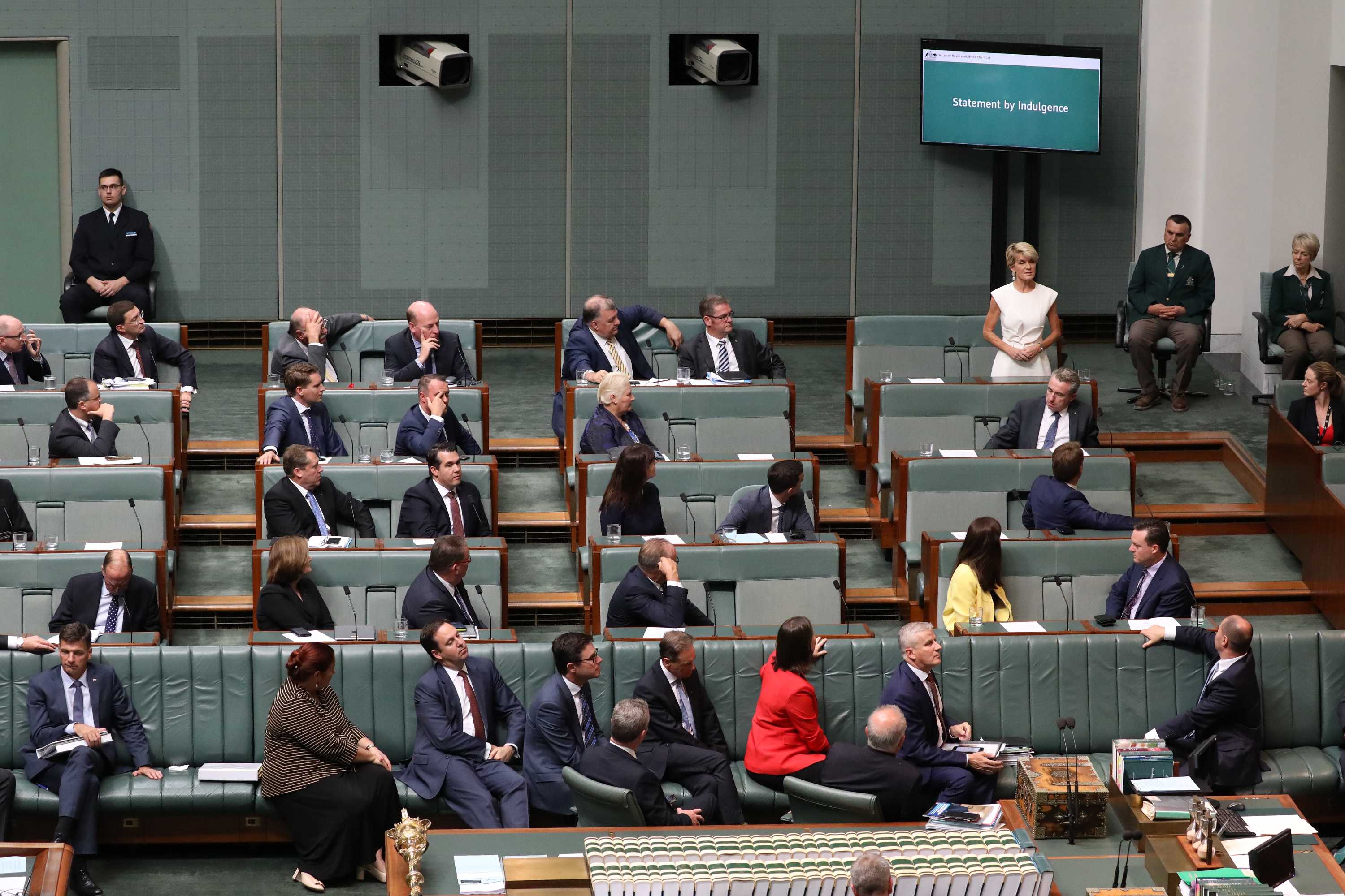 Julie Bishop, standing and wearing a white dress, speaks in the House of Representatives as other MPs watch on.