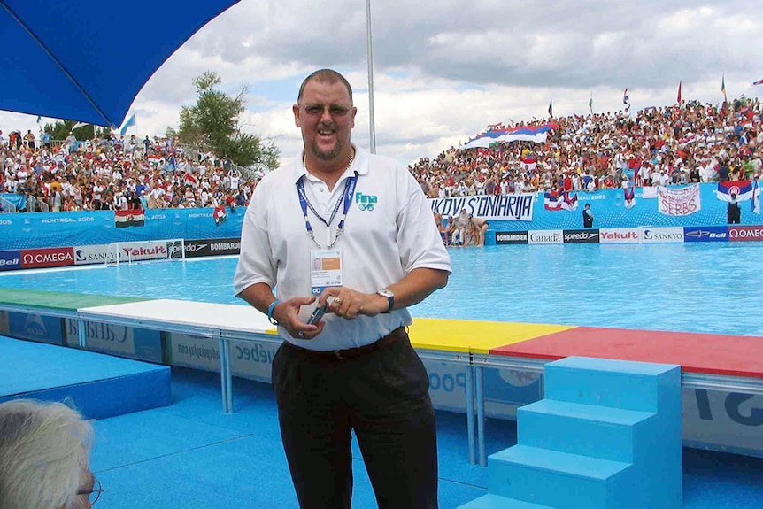 Man in white shirt stands in front of pool displaying his Olympic accreditation pass. 