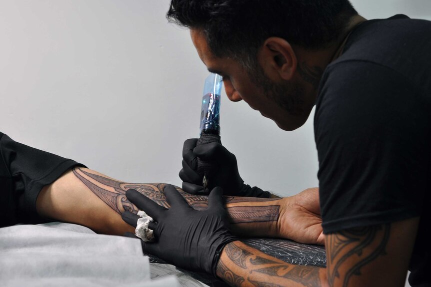 A Maori man focuses as he tattoos a traditional Moko design onto the arm of a man.