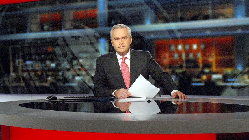 Huw Edwards holding a sheet of paper sitting at a news desk 