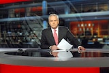 Huw Edwards holding a sheet of paper sitting at a news desk 