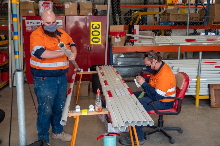 two men in hi-vis tape reflectors to poles in a workshop shed