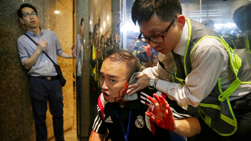 A man raises a bloody left hand as someone in a high-vis vest clasps a towel to the man's left ear which is severely wounded.