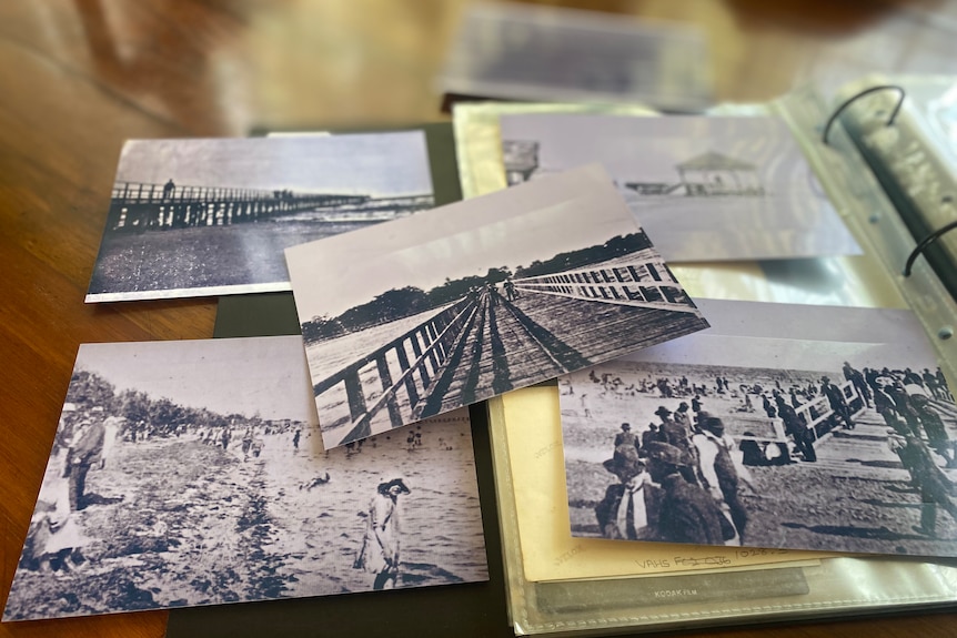 Old black and white pictures of Altona Pier are seen stacked on top of each other on a wooden table.