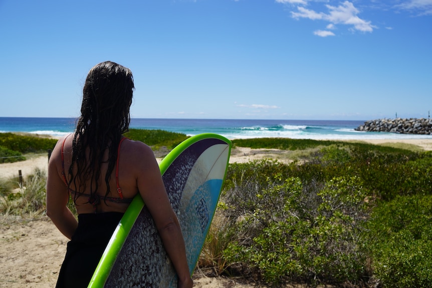 A woman looks out in to the ocean at waves breaking, holding a green surfboard.