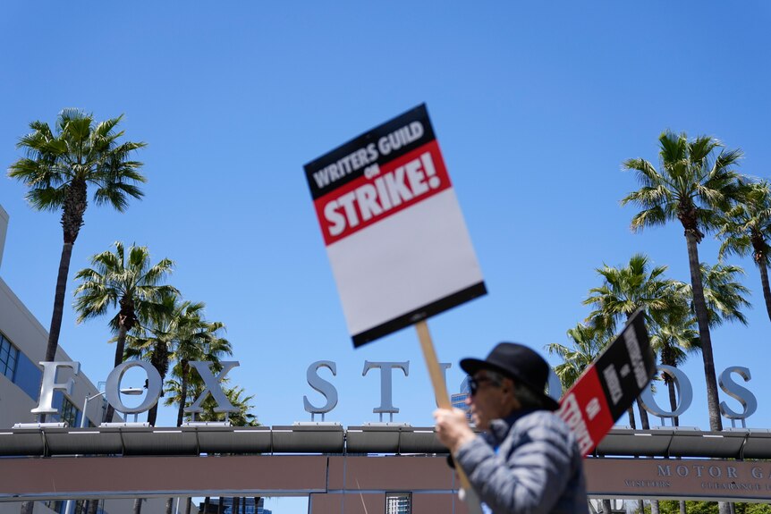 A man holding a sign with Writer's Guild Strike walks oustide a signs displaying Fox Studios.