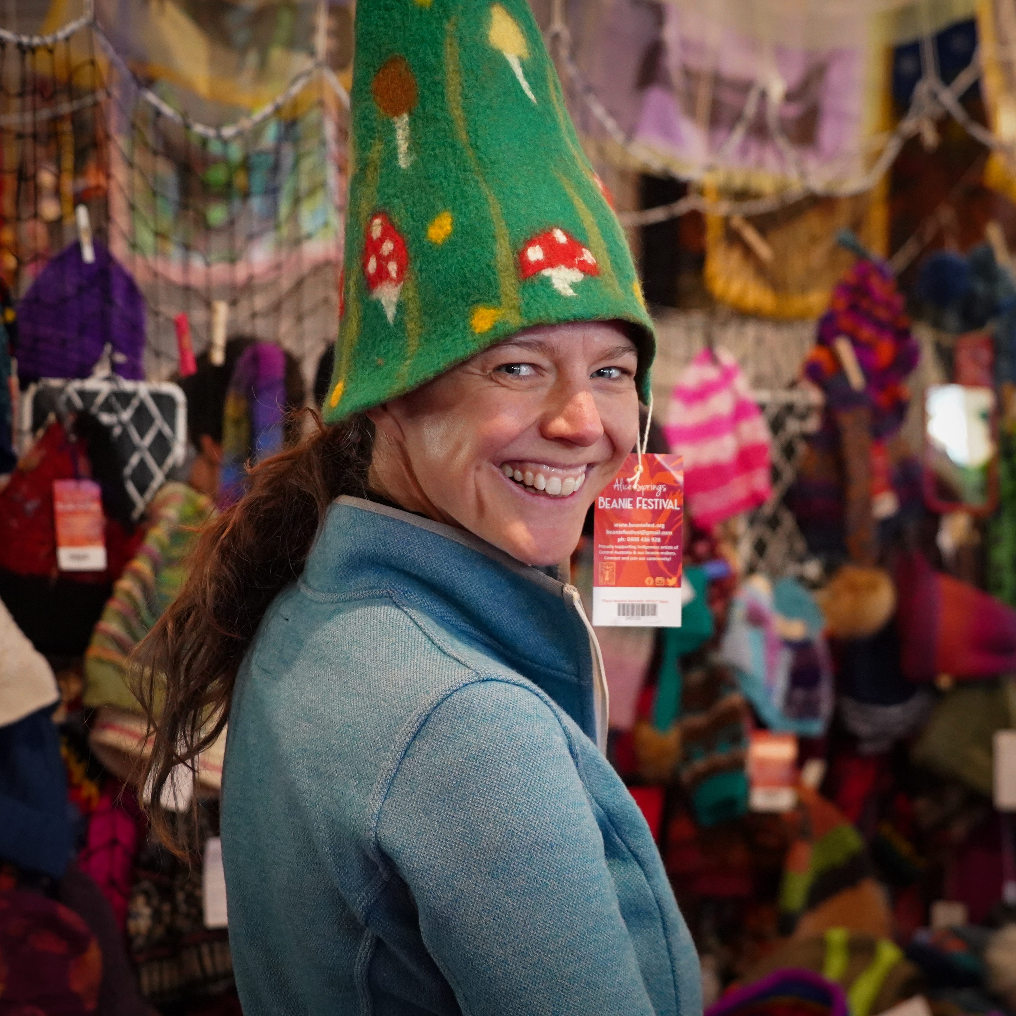 A woman smiling at the camera while wearing a tall green felt hat, red and white mushrooms are dotted across the beanie.