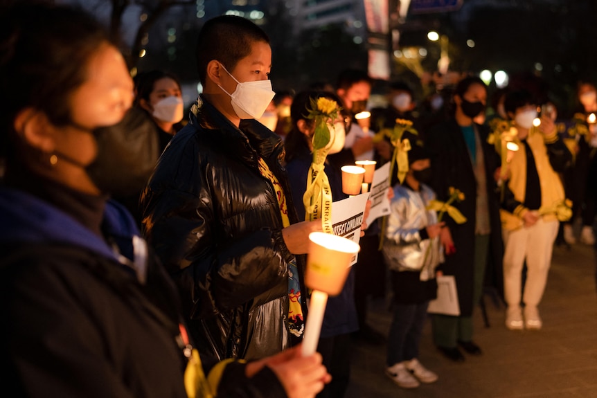 A crowd of people gathered at night hold small candles and placards.