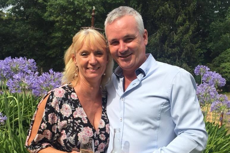 A woman holding a glass of wine and man holding a beer stand together smiling in front of purple flowers (agapanthus) and trees.