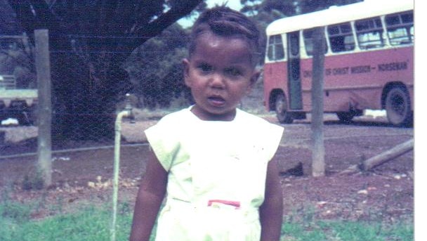 An old, colour photograph of a toddler standing outside. A bus is visible in the background.