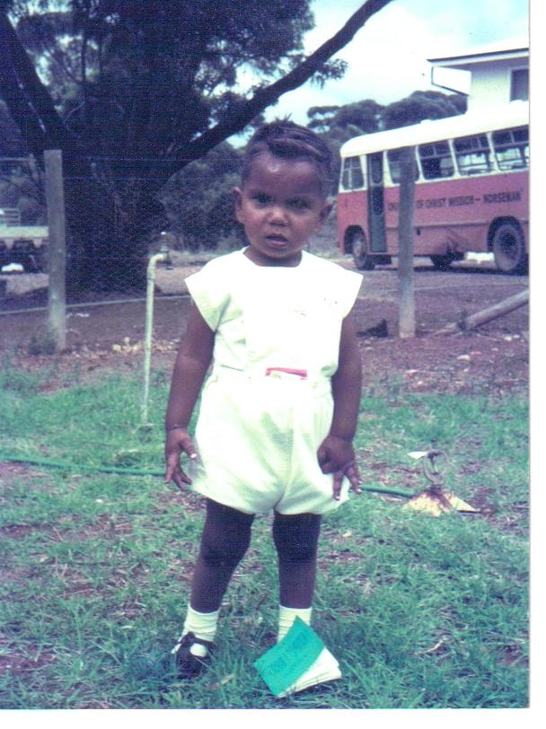 An old, colour photograph of a toddler standing outside. A bus is visible in the background.