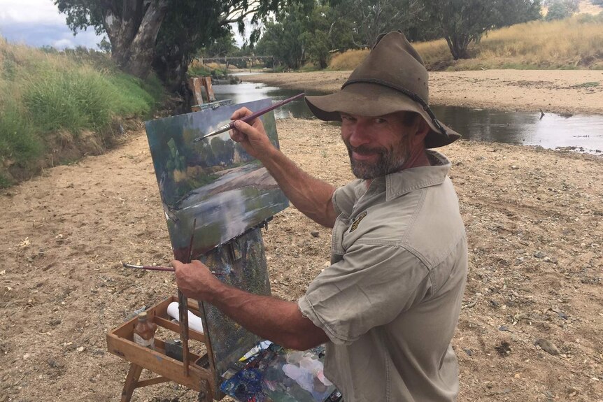 Brett Garling poised with his paint brush at his easel on the banks of a small river.