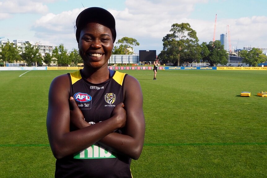 A woman dressed in a AFLW uniform stands with arms crossed and smiling looking at the camera on a football oval.