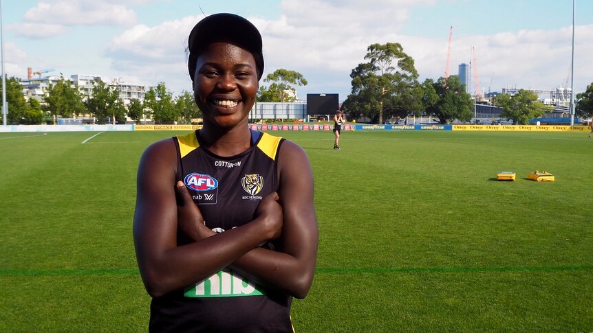 A woman dressed in a AFLW uniform stands with arms crossed and smiling looking at the camera on a football oval.